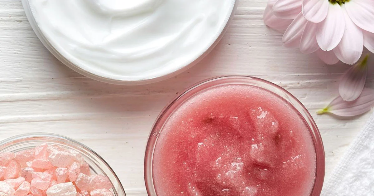 Top view of spa products including pink scrub, lotion, pink salts, and flower on a white surface.
